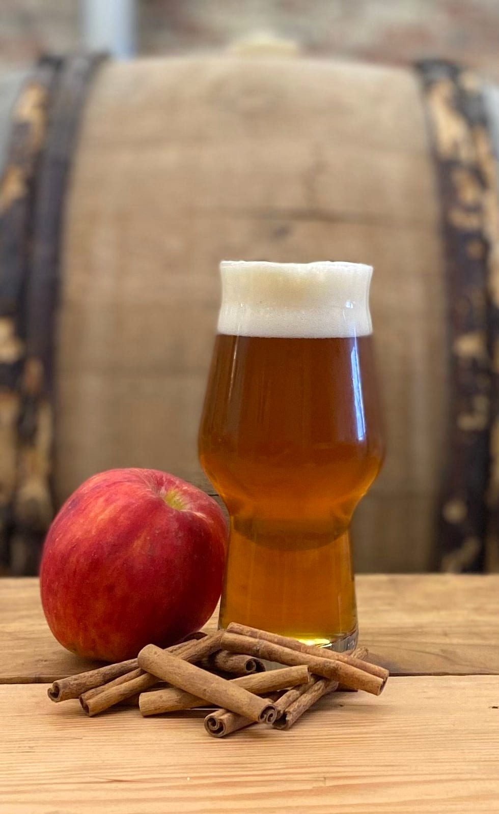 A glass of amber beer beside a red apple and cinnamon sticks in front of a barrel