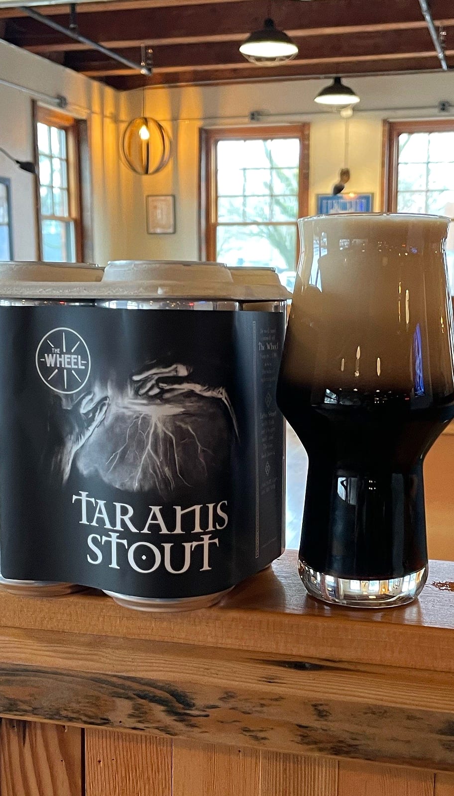 A four-pack of stout cans next to a glass of dark beer on a wooden ledge inside the pub