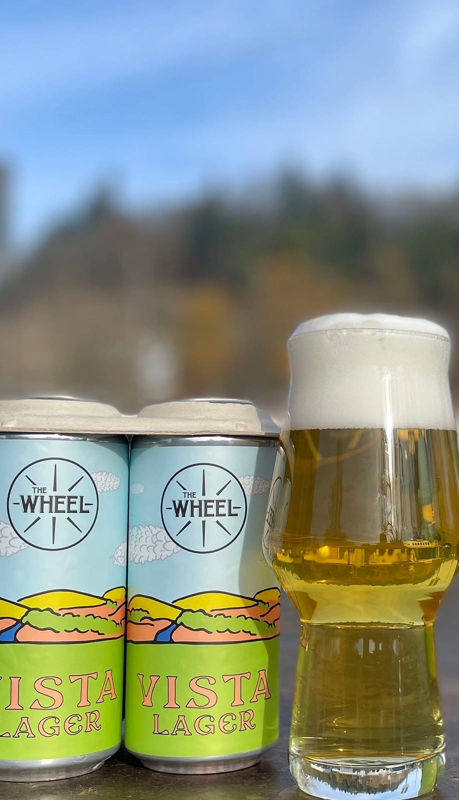 A four-pack of lager cans beside a glass of pale beer outdoors with hills behind