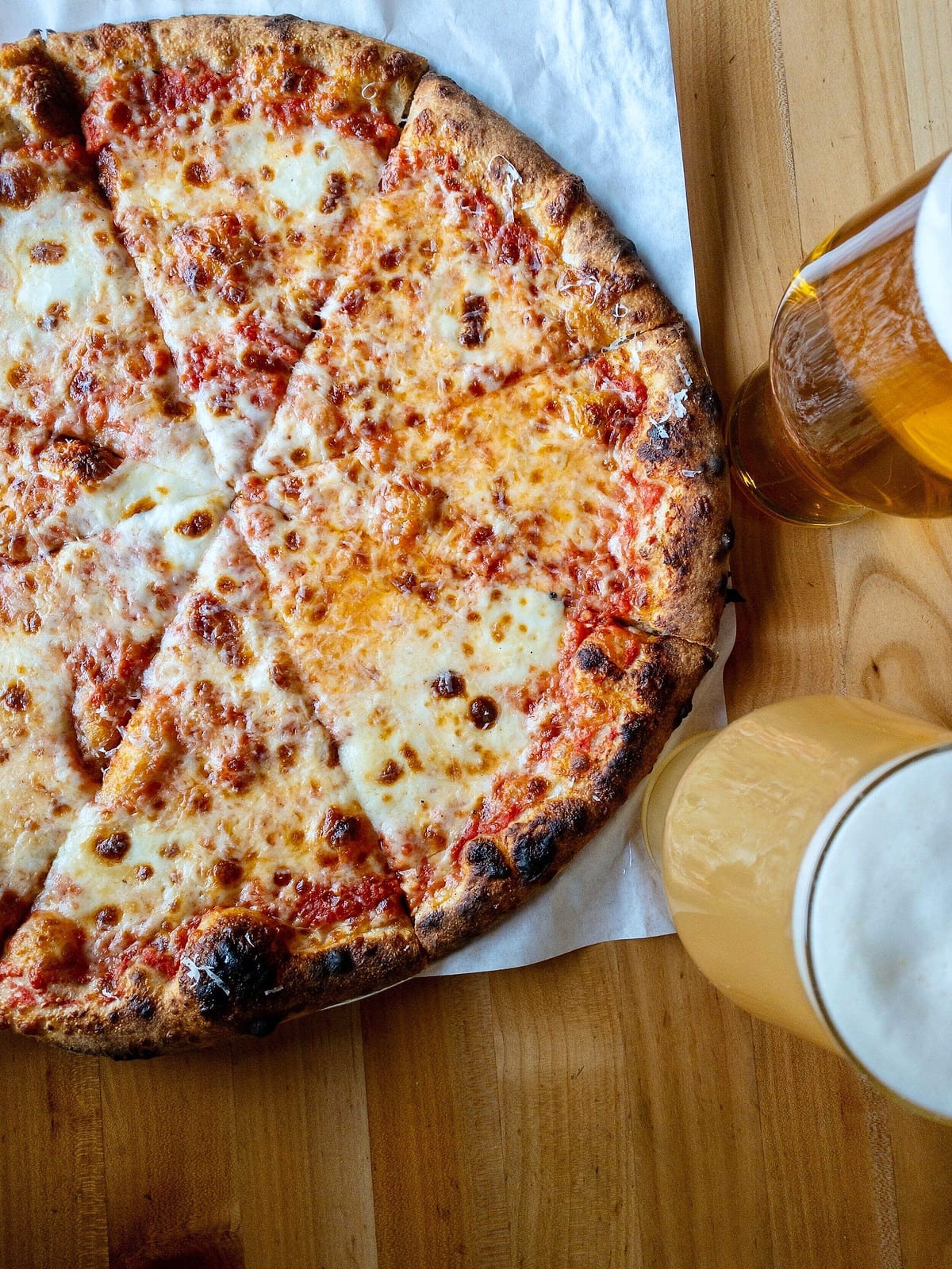 Overhead view of a cheese pizza on parchment beside two glasses of beer on a wood table