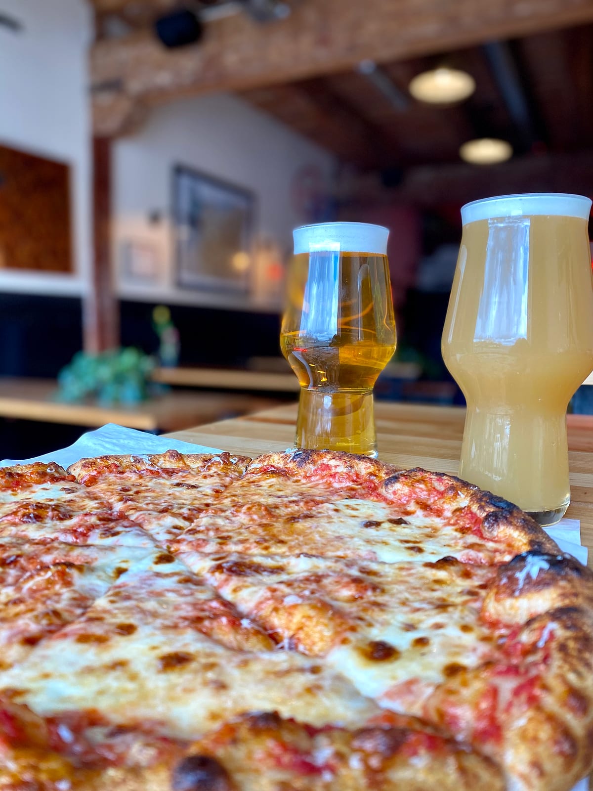 Close-up of a cheese pizza with two glasses of beer and the dining room behind